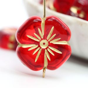 May include: Close-up of a red glass bead with a flower design. The bead has a gold-colored floral pattern and is strung on a gold-colored wire. The bead is in focus, with other beads and a white bowl in the background.