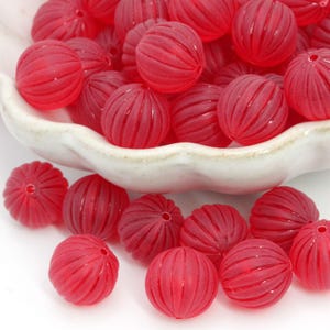 May include: A collection of translucent red beads with a ribbed, spherical design. The beads are scattered in a white ceramic bowl. These decorative beads are suitable for jewelry making and craft projects.