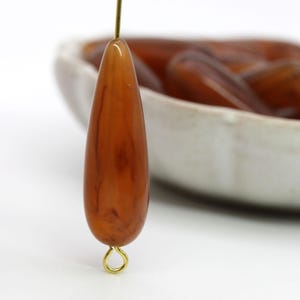 May include: A single brown teardrop-shaped glass bead with a gold jump ring attached. The bead has a marbled pattern and is sitting on a white surface.