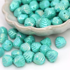 May include: A collection of turquoise-colored beads with a textured, ridged design. The beads are scattered, some spilling out of a white, scalloped-edge bowl. The beads have a glossy finish and a hole for stringing.