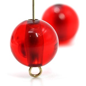 May include: Two translucent red round beads with a gold-colored eye pin. One bead is in focus, showing vertical lines within the red sphere. The second bead is out of focus in the background. The beads are likely for jewelry making.
