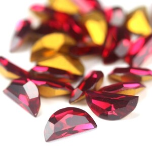 May include: A collection of faceted, half-moon shaped, red rhinestones with a metallic gold backing. The gemstones are scattered across a white surface, with some in focus and others blurred.