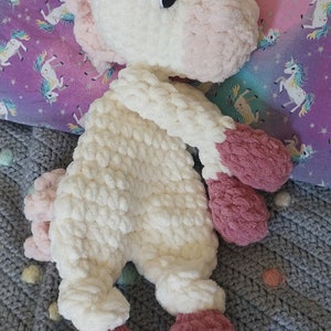May include: A white and pink crocheted unicorn plush toy with a curled horn. The toy is lying on a gray and white crocheted blanket.