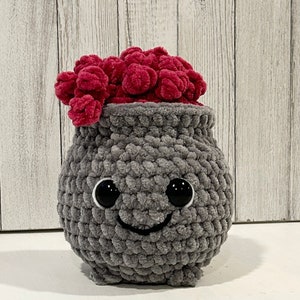 May include: A gray crocheted cauldron with a smiling face and large black eyes. The cauldron is filled with a pile of bright pink yarn.