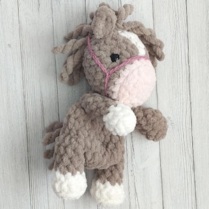 Op de afbeelding: Een gehaakt pluche speelgoedpaard met bruin en wit bont, met een roze gehaakt halster. Het paard heeft een wit gezicht met zwarte ogen en een roze neus.