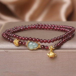 May include: A double-strand bracelet featuring small, round, deep red garnet beads. The bracelet includes a central pale green, gourd-shaped stone set in gold, a textured gold bead, and a small, detailed gold teapot charm hanging from the strand.