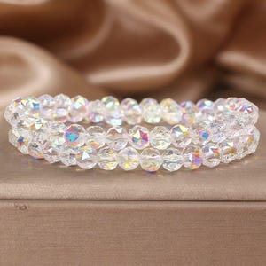 May include: A two-strand bracelet made of clear, faceted beads. The beads have an iridescent sheen, reflecting various colours. The bracelet is presented on a neutral surface, with a soft, blurred background.