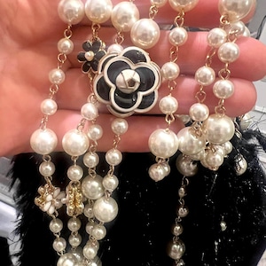 Puede incluir: Un collar de perlas con un colgante de flor en blanco y negro y pequeños detalles florales. El collar presenta múltiples hilos de perlas de imitación de varios tamaños, conectados por cadenas doradas. El colgante de flor negra tiene un centro plateado.