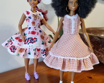 Könnte beinhalten: Zwei Puppen, die Kleider tragen. Die Puppe links trägt ein rosa-weißes Kleid mit einem Kirsch- und Beerendruck. Die Puppe rechts trägt ein rosa-weißes Gingham-Kleid mit einer weißen Spitzenborte.