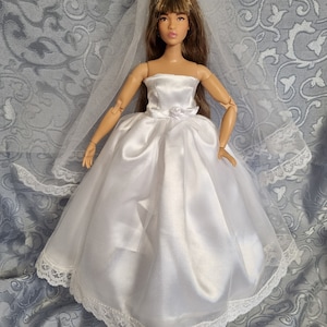 May include: A doll wearing a white satin wedding dress with a tulle overlay and lace trim. The strapless gown features a small bow at the waist. A white veil with lace edging and a headpiece complete the bridal look.