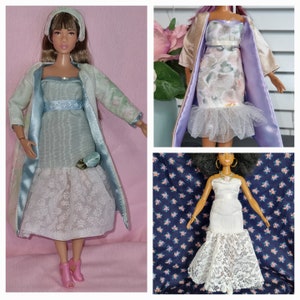 Könnte beinhalten: Drei Puppen, die verschiedene Kleider tragen. Die Puppe links trägt ein hellblaues Kleid mit einer weißen Spitzenüberlage und eine hellgrüne Jacke. Die Puppe in der Mitte trägt ein Kleid mit Blumenmuster mit einer weißen Spitzenüberlage und eine lila Jacke. Die Puppe rechts trägt ein weißes Kleid mit einer weißen Spitzenüberlage.