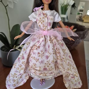 以下が含まれることがあります： 白いシャツとピンクの花柄のドレスにピンクのリボンをつけた人形。人形は茶色の髪と茶色の目です。