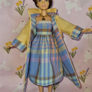 May include: A doll wearing a yellow jacket over a plaid dress with a white lace trim. The doll has brown hair and is wearing white shoes.