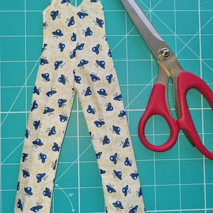 May include: A pair of yellow and blue patterned overalls with a V-neck and thin straps. The overalls are laid out on a green cutting mat with a pair of red handled scissors.
