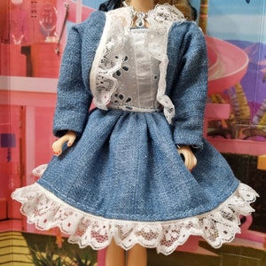 Könnte beinhalten: Ein Jeanskleid mit weißem Spitzenbesatz und einer passenden Jeansjacke. Das Kleid hat ein weißes Spitzenoberteil mit Blumenmuster. Die Puppe trägt eine silberne Halskette.