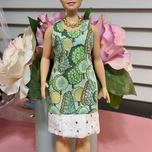 May include: A doll wearing a green dress with a white lace trim and a red headband. The doll has blonde hair and is sitting on a light blue planter with pink flowers.