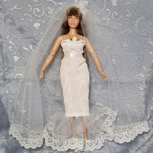 Könnte beinhalten: Ein weißes Brautkleid mit einer Spitzenborte und einer weißen Rose am Dekolleté. Die Puppe trägt einen weißen Schleier und eine Tiara.