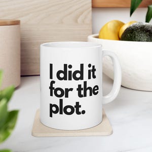 May include: White ceramic mug with the text "I did it for the plot" in black, on a beige coaster. The mug has a curved handle. A blurred background includes a bowl of fruit and wooden surfaces.