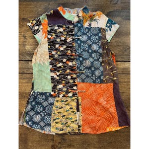 May include: A sleeveless top featuring a patchwork design of various fabrics in orange, blue, black, yellow, and cream. The garment has a mandarin collar, a front opening with five fabric-covered toggle closures, and a mix of floral and geometric patterns.