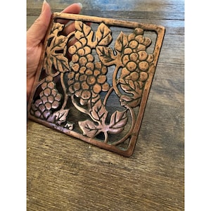 May include: A square copper-toned metal trivet featuring an openwork design of grape clusters and vine leaves. The metallic surface displays a warm, burnished finish with intricate detailing of the fruit and foliage across the entire frame.