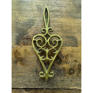 Pode incluir: Um descanso de panela em metal dourado com um design vazado simétrico e alongado, apresentando volutas intrincadas e uma alça superior pontiaguda para pendurar. A peça possui um acabamento polido e um formato distinto de coração.