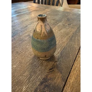 May include: A small, teardrop-shaped ceramic vase with a narrow neck and a wide base. The surface features a mottled brown and tan glaze with a prominent horizontal band of muted blue-green color encircling the middle and textured vertical ridges.