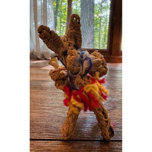 May include: A handcrafted straw figurine shaped like a donkey, featuring woven tan fibers, a small blue yarn accent near the head, and a skirt made of bright red and yellow yarn strands wrapped around the midsection of the animal-shaped structure.
