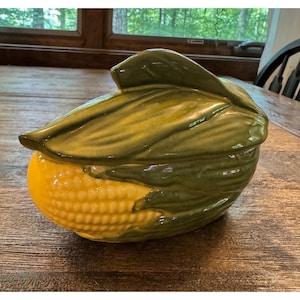 May include: A ceramic lidded serving dish shaped like an ear of corn. The base features a bright yellow textured surface representing corn kernels, while the removable lid is moulded to look like dark green corn husks with a glossy finish.