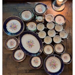 May include: A large collection of white porcelain dinnerware featuring a dark blue border with gold trim and a central floral bouquet pattern. The set includes various plates, bowls, teacups, saucers, a lidded sugar bowl, and a large oval platter.