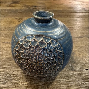 May include: A round ceramic vase with a deep blue glaze and a textured, dark brown circular relief pattern on the front. The vase features a narrow neck with a thick rim and horizontal ridges around the upper body, showcasing a handcrafted finish.