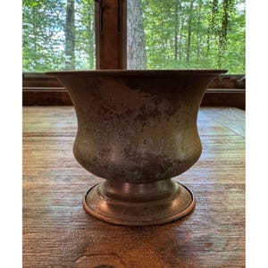 May include: A weathered, dark metallic urn-shaped planter with a wide, flared rim and a circular pedestal base. The exterior surface displays a mottled, oxidized patina with shades of brown, bronze, and dark gray, showing signs of age and wear.
