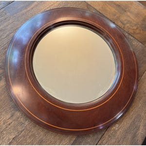 May include: A circular wall mirror featuring a dark brown wooden frame with a smooth, polished finish. The frame is accented by a thin, lighter-colored circular inlay line that runs along the inner portion of the wood, surrounding the glass.