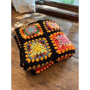 May include: A folded crochet blanket featuring a pattern of multi-colored granny squares. Each square is bordered by thick black yarn, with interior colors including bright yellow, pink, orange, green, and blue in a repeating geometric design.