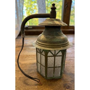 May include: A wall-mounted lantern with a weathered green metal finish and a curved arm. The fixture features a hexagonal body with textured, pebbled glass panels and a tiered top cap. A black electrical cord extends from the base.