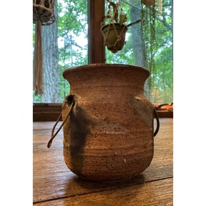 May include: A rustic, cylindrical ceramic pot with a textured, speckled brown finish. The vessel features a wide, flared rim and two side-mounted metal wire handles. Dark, charcoal-colored glaze accents run vertically down the sides of the pot.
