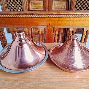May include: Two copper tagine pots with lids. One pot has a smooth, reflective surface, while the other has a textured, hammered design. Both sit on a wooden table, with a wooden background.