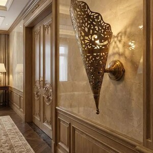May include: A wall-mounted sconce featuring a conical, brass-toned metal shade with intricate, laser-cut filigree patterns. The fixture is attached to a circular base plate and emits a warm, soft glow through the detailed cutouts in the metal.