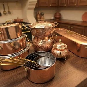 Puede incluir: Una colección de utensilios de cocina de cobre que incluye una pila de tres ollas redondas con tapas, una fuente de horno ovalada grande con tapa, un cuenco pequeño con tapa y un juego de cacerolas encajables con mangos largos de latón.