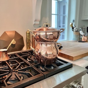 Op de afbeelding: Een tweekoppige gestoomde koperen stoompan met messing handgrepen en een bijpassend deksel, rustend op een zwart gasfornuis. Het kookgerei heeft een gepolijste metalen afwerking met een getextureerd oppervlak en een rond, stapelbaar ontwerp.