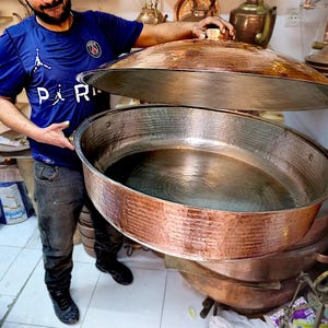 Può includere: Una grande pentola rotonda in rame martellato con coperchio a cupola abbinato. La superficie esterna presenta una finitura testurizzata e punteggiata, mentre l'interno è liscio e metallico. La pentola ha un bacino ampio e aperto.