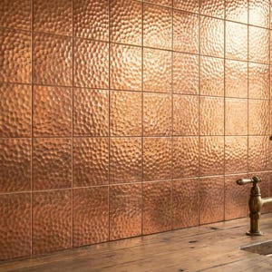 Puede incluir: Una pared cubierta con azulejos de cobre martillado cuadrados con un acabado metálico reflectante. Un grifo de puente de tono latón con dos manijas está montado cerca del borde de una encimera de madera, junto a un fregadero revestido de cobre.