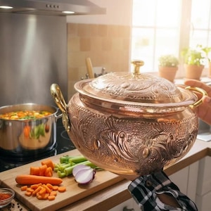 May include: A round, polished copper pot with a matching lid, featuring intricate embossed floral and leaf patterns across the surface. The pot includes two curved gold-toned metal handles on the sides and a small, round gold-toned knob on the lid.