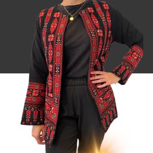 May include: Black jacket with detailed red embroidery on the front and sleeves. The jacket has a floral and geometric pattern. The sleeves are long and flared. The jacket is worn over a black top and trousers.