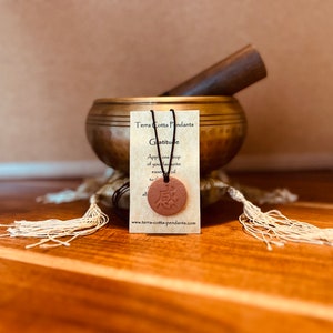 Puede incluir: Colgante de terracota con la palabra "Gratitud" escrita en caracteres japoneses en un disco de terracota marrón. El colgante está en una tarjeta con el texto "Terra Cotta Pendants Gratitude Apply one drop of your favourite essential oil to the disc... www.terra-cotta-pendants.com"