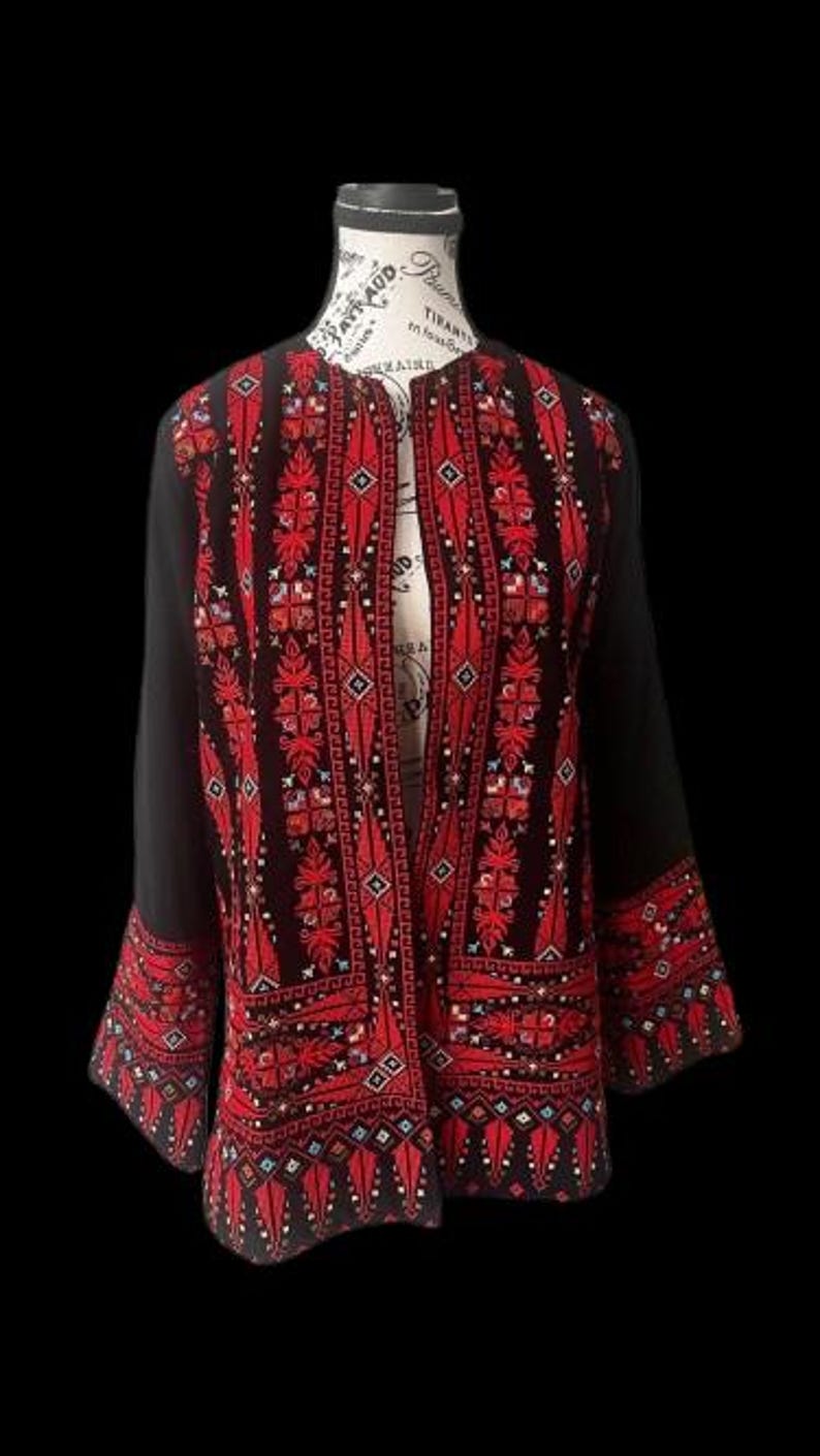 May include: Black jacket with detailed red embroidery. The open-front jacket showcases intricate patterns on the front, sleeves, and cuffs. The embroidery incorporates geometric and floral designs, providing a striking contrast against the black fabric. The jacket is displayed on a mannequin.