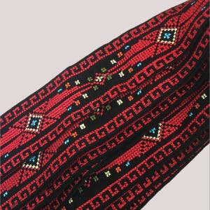 May include: Close-up of a textile with a repeating geometric pattern. The fabric is primarily black with red, blue, and white embroidered details. The design features a series of squares and lines.