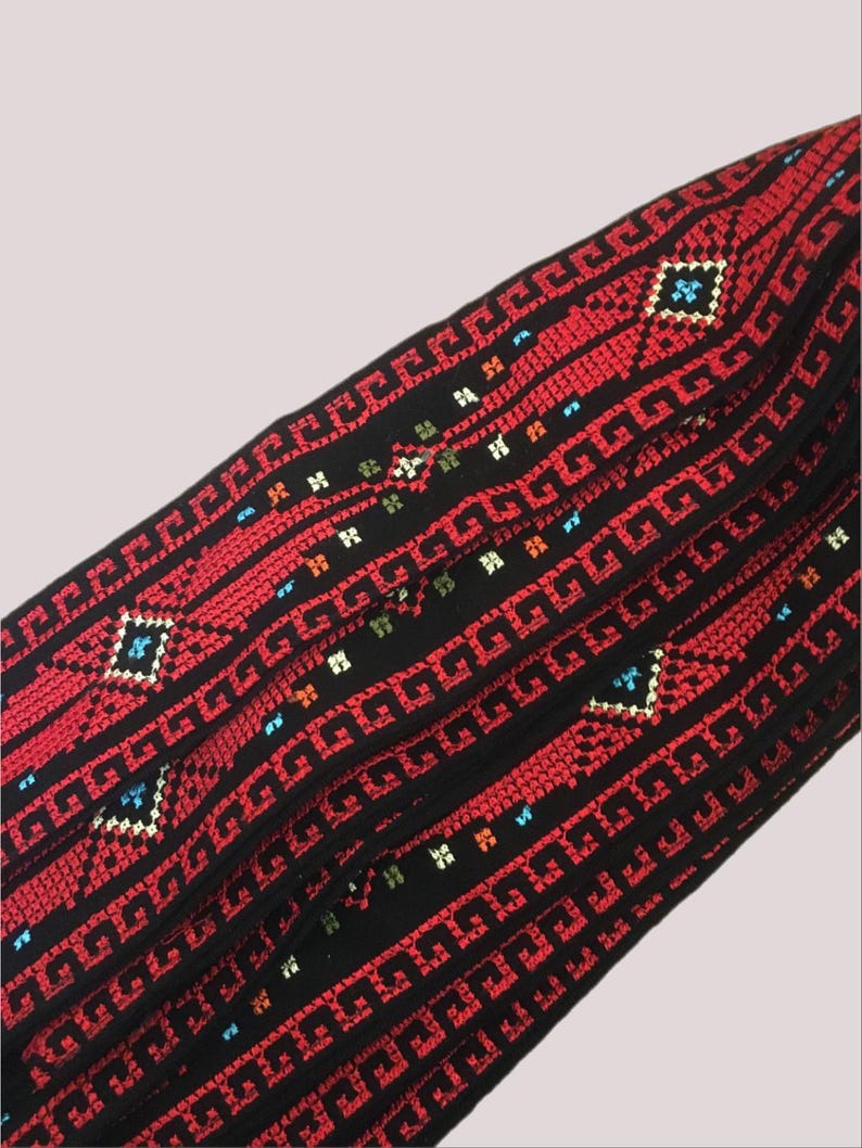 May include: Close-up of a textile with a repeating geometric pattern. The fabric is primarily black with red, blue, and white embroidered details. The design features a series of squares and lines.