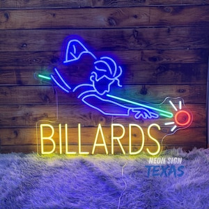 May include: A neon sign featuring a blue outline of a person playing billiards with a green cue stick and a red ball. Below the figure, the word BILLARDS is illuminated in yellow neon light. The sign is mounted on a dark wooden plank wall.