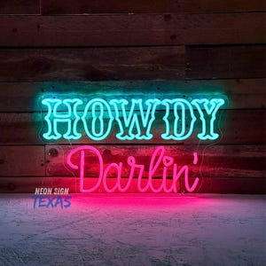 以下が含まれることがあります： ターコイズブルーとピンクのカラーリングで「Howdy Darlin'」という文字が書かれたネオンサイン。サインは木製の壁に取り付けられています。