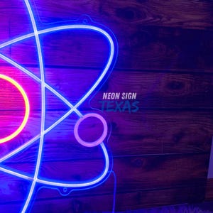 May include: A neon sign in the shape of an atom, with a blue outline and white internal lines. The center is a bright pink and yellow circle. The text "NEON SIGN TEXAS" is in the lower right corner.